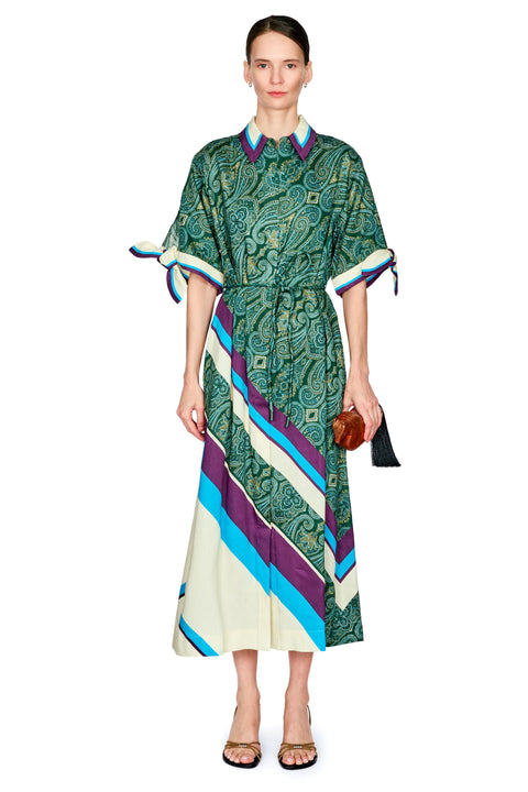 A woman wearing a long-sleeved midi dress with a green paisley pattern and bold diagonal stripes in cream, blue, and purple. The dress features a pointed collar and tie details on the sleeves.