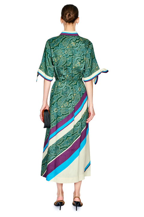 A woman viewed from the back wearing a midi dress with a green paisley pattern and diagonal stripes in cream, purple, and blue. She is wearing black pointed-toe heels and holding a black clutch against a plain white background.