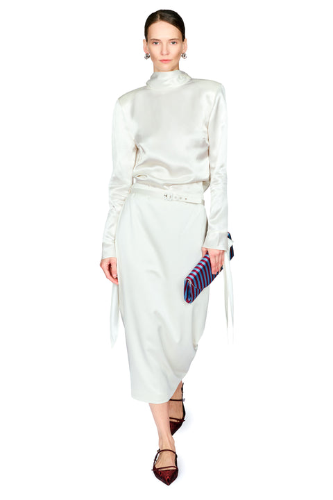 A model wearing a white satin long-sleeve mock-neck dress with a matching belt, holding a striped clutch, on a solid white background.