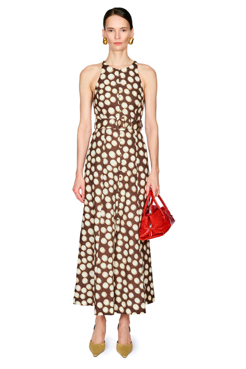 A model stands against a white background wearing a brown halter-neck maxi dress with a cream polka dot pattern, holding a small red handbag.