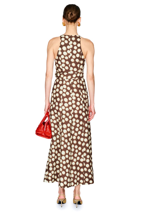 A woman shown from the back wearing a sleeveless halter-neck maxi dress with a brown and beige polka dot pattern. She is holding a small red handbag and wearing gold pointed-toe heels against a plain white background.