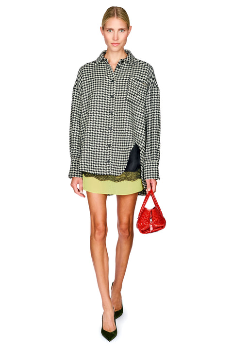 A model wearing a black and white houndstooth button-up shirt, a light olive green mini skirt, and holding a red handbag against a plain white background.