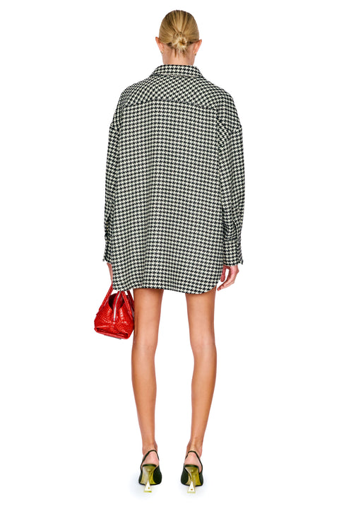 A model shown from the back wearing an oversized black and white houndstooth shirt, holding a small red handbag and wearing green and gold high heels against a white background.