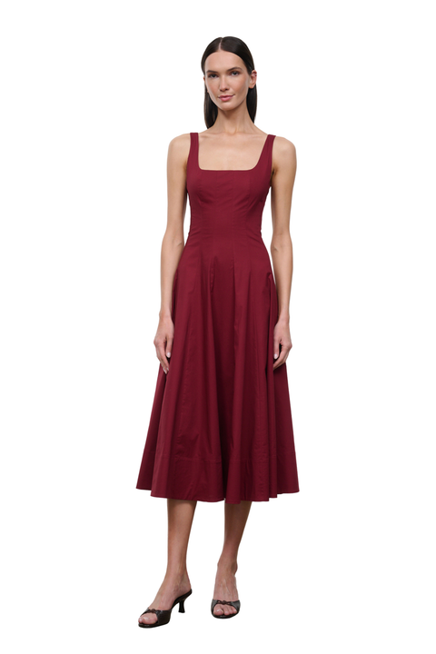 A woman modeling a sleeveless midi dress in deep burgundy with a square neckline and a flared skirt, paired with black heeled sandals, against a plain white background.